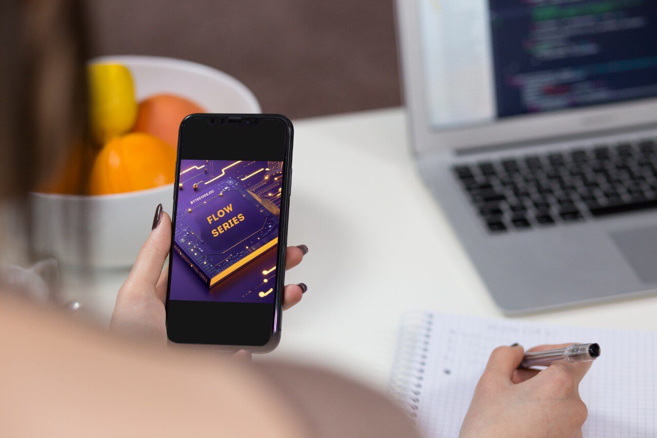 Person holding a phone with a 'Flow Series' course open, sitting at a desk with a laptop and fruit bowl.